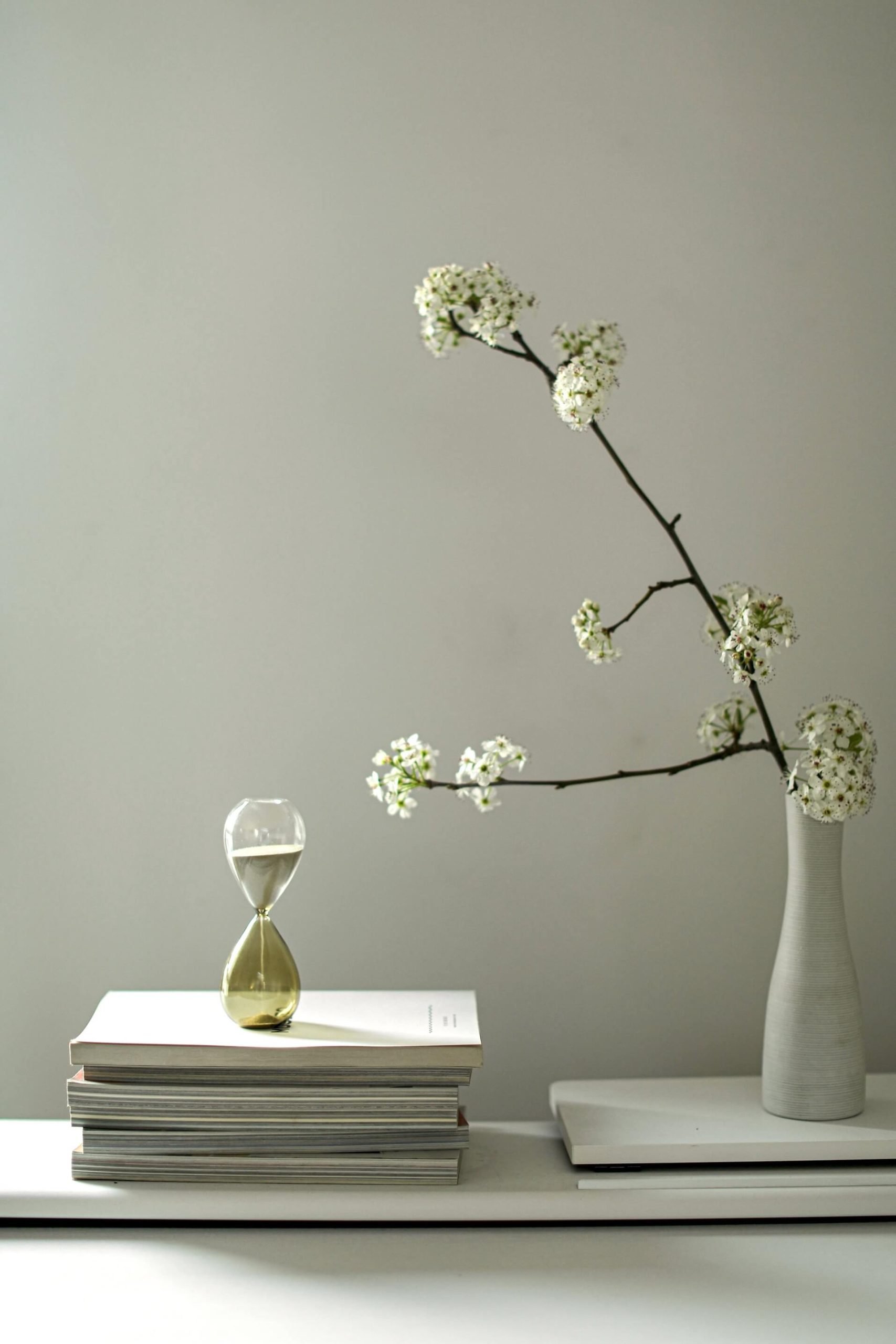 a quiet workspace with books, an hourglass and a vase with branches in natural light, reflecting a moment to pause and a gift to yourself